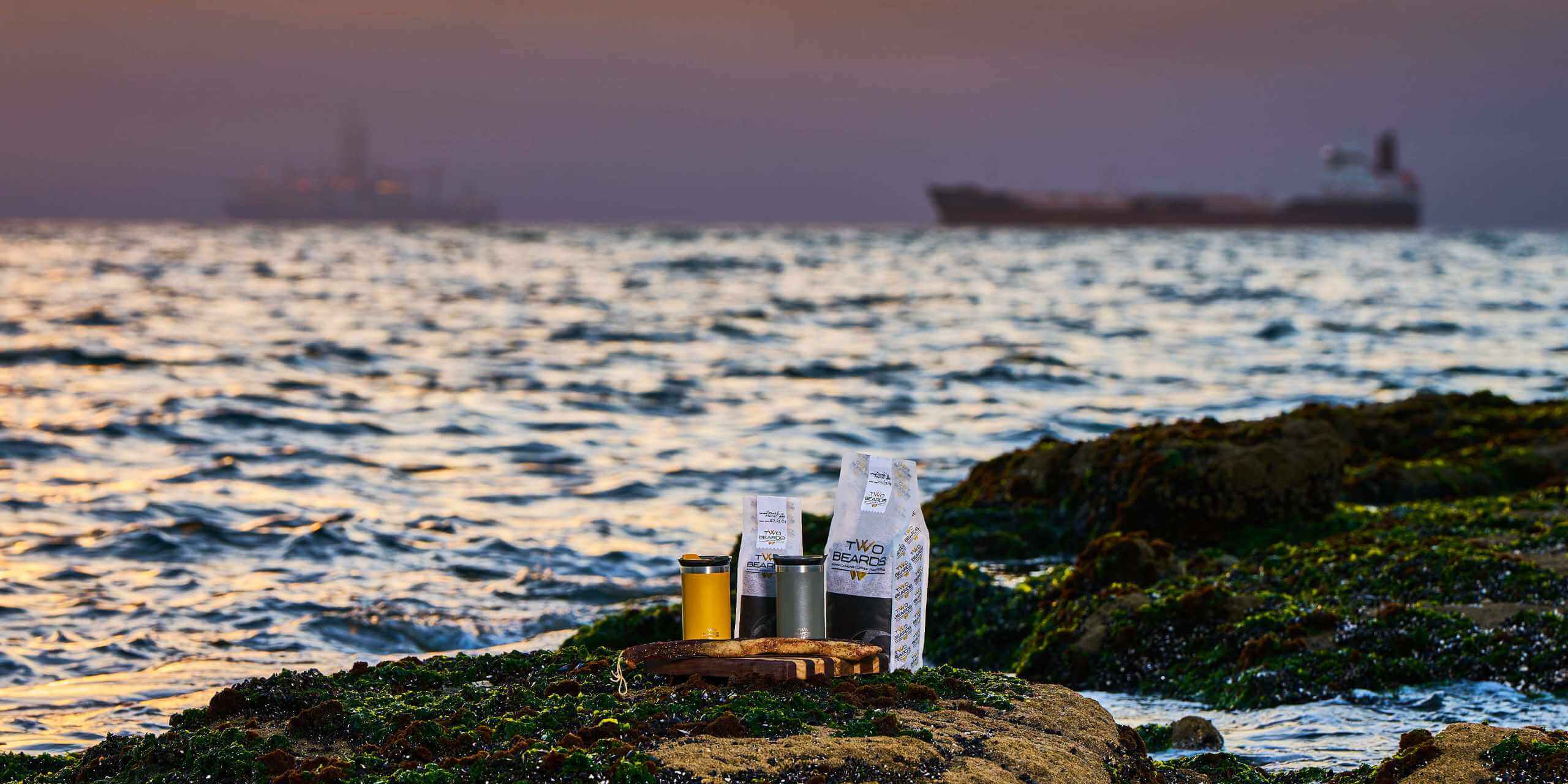 Two Beards Coffee by the Namibian Coast: The image showcases Two Beards Coffee set against the serene backdrop of Namibia's coastline. The calm sea and a distant ship are silhouetted against the soft hues of the sunset, while two coffee cups rest on a moss-covered rock, inviting viewers to enjoy a peaceful coffee moment by the sea.