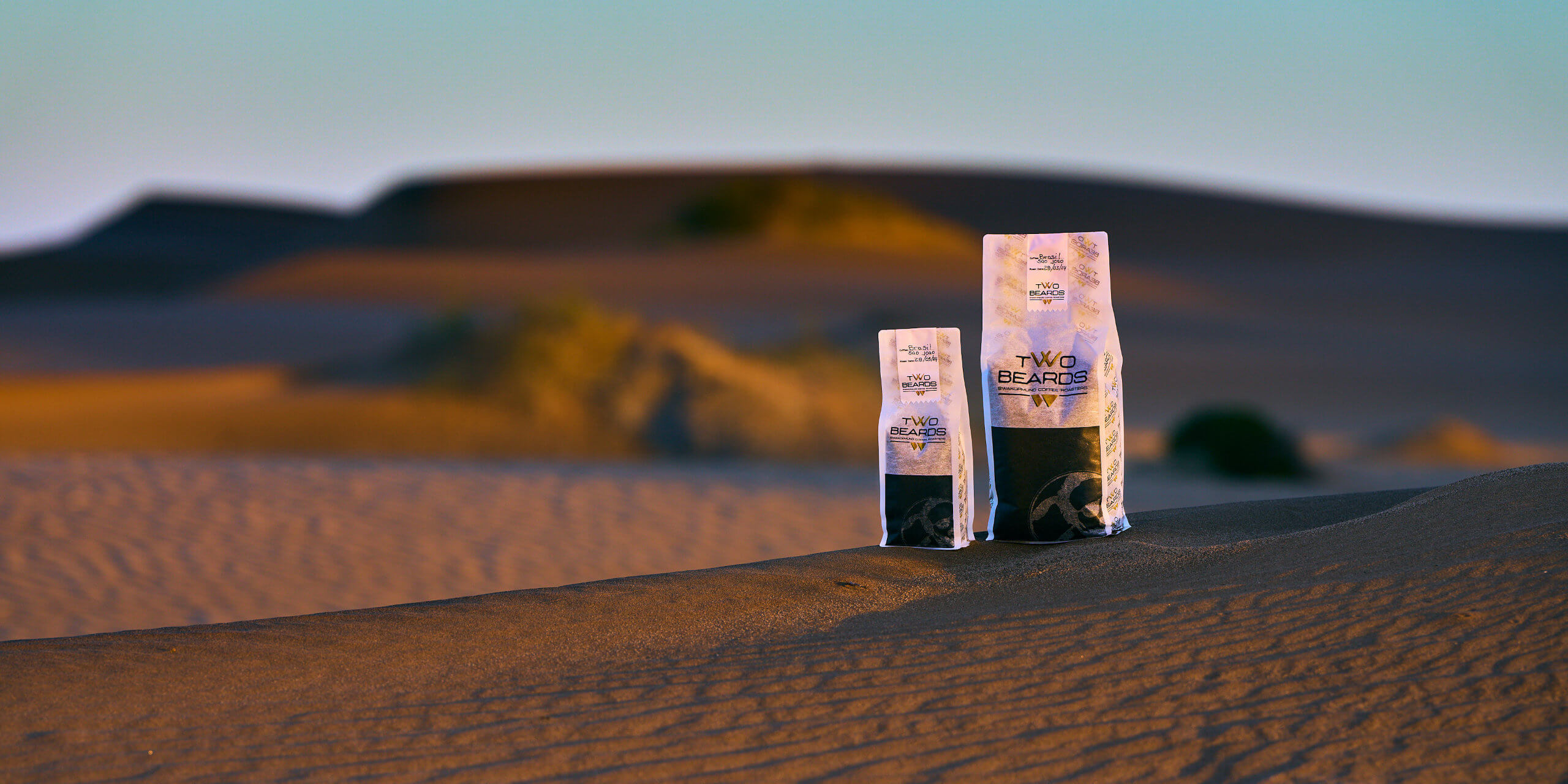 Beard Coffee Bags in Namib Dunes: The photo captures two Beard Coffee Bags placed amidst the vast dunes of the Namib Desert. The bags are prominently featured against the backdrop of the rolling sandy landscape, highlighting the contrast between the product and the natural beauty of the desert.
